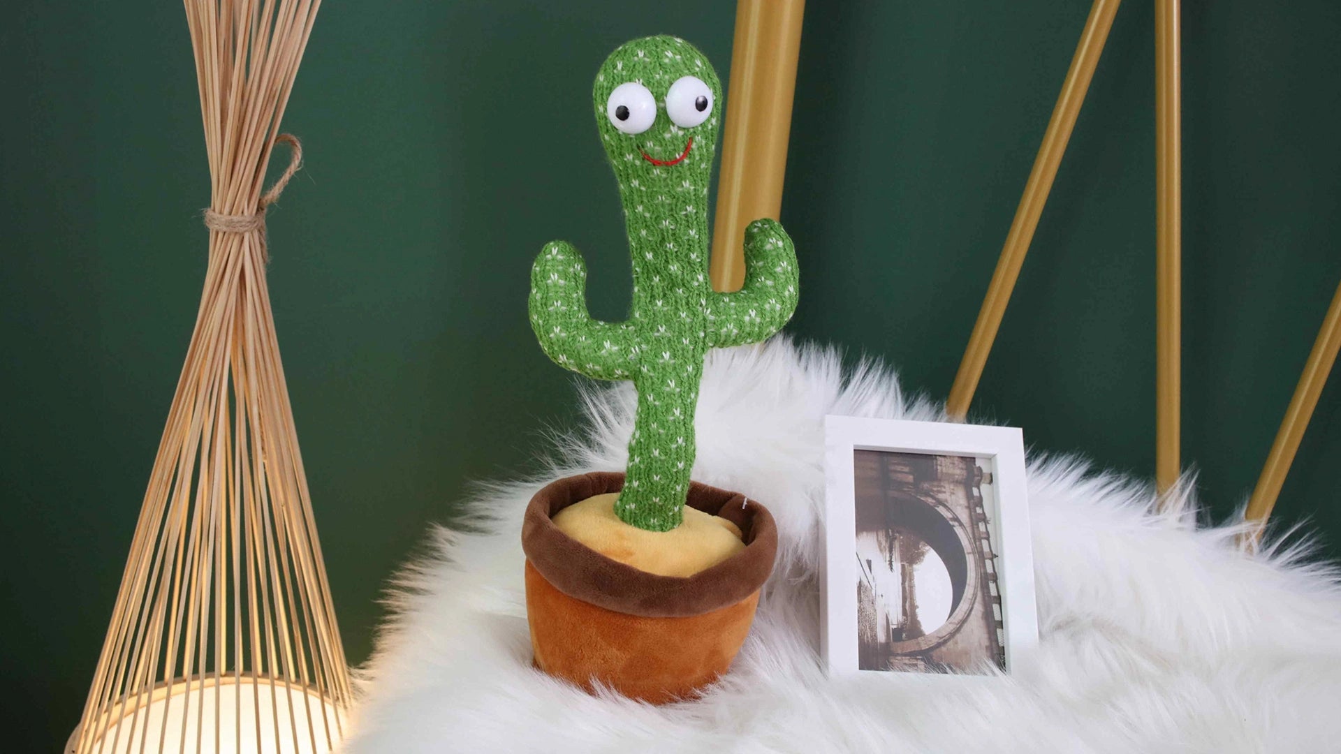 green background, white feather blanket, photograph frame, yellow bamboo stick lamp, brown flowerpot, green singing dancing cactus, dancing cactus toy, voice activated cactus, talkback speaking cactus, talking baby cactus toy, white big eyes,